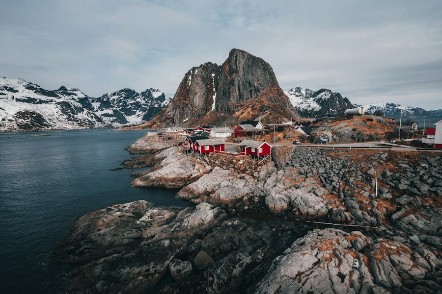Red wooden huts on a rocky shoreline with mountains beyond